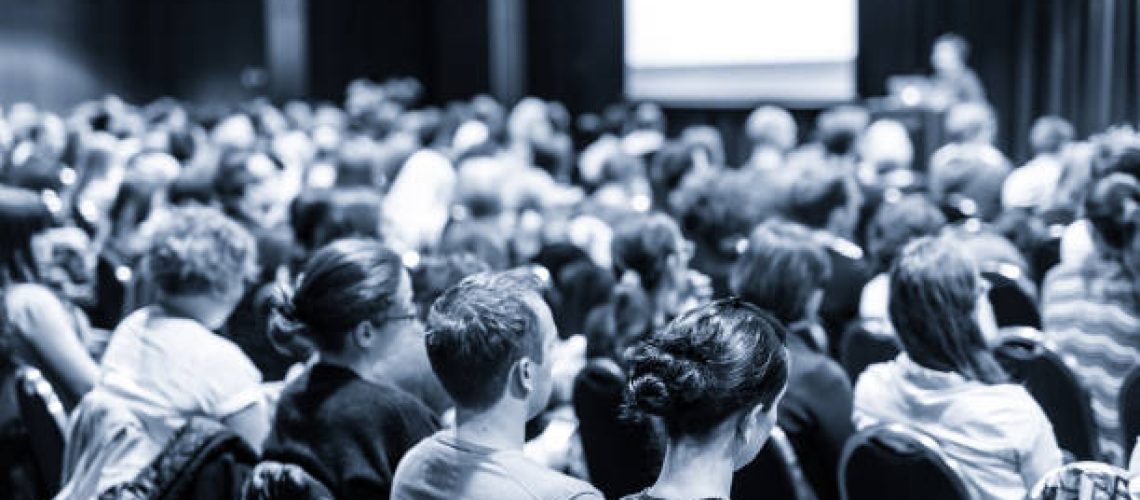 Business and entrepreneurship symposium. Speaker giving a talk at business meeting. Audience in conference hall. Rear view of unrecognized participant in audience.